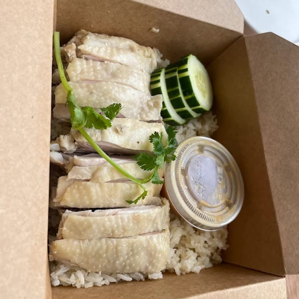 Hainanese chicken rice with sliced chicken, cucumber, cilantro, and sauce in a takeout box.