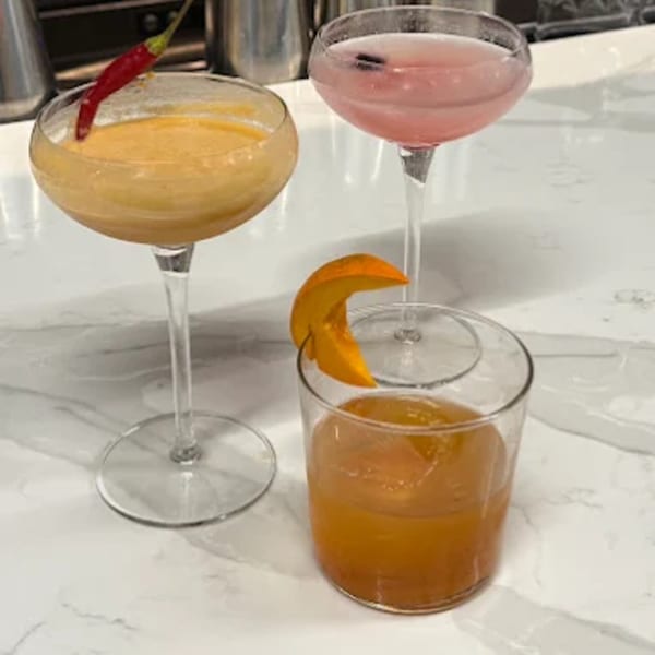 Three Thai-inspired cocktails: one yellow with a red chili, one pink, and one orange with a fruit garnish.