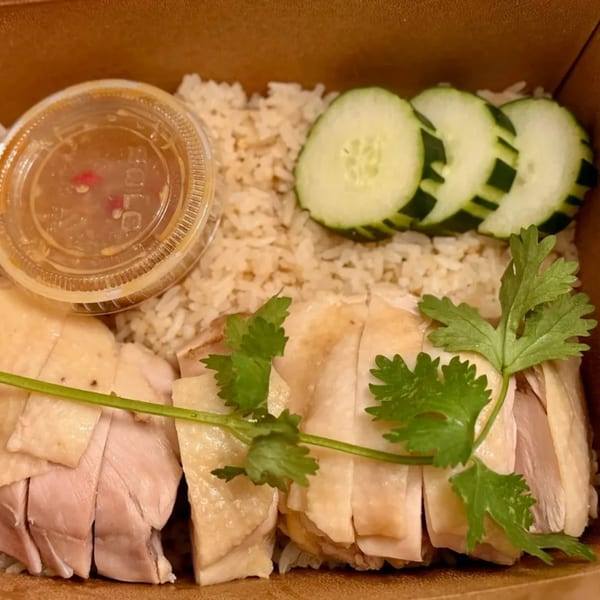 Thai Hainanese chicken rice with sliced chicken, rice, cucumbers, cilantro, and sauce in a takeout box.