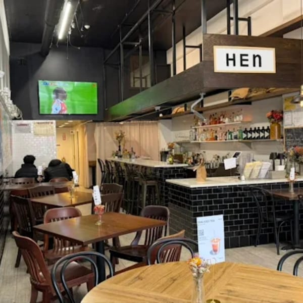 Interior of Hen restaurant with wooden tables, black chairs, a bar, and a TV showing sports.