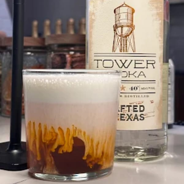 A layered Thai iced tea cocktail with white foam, amber liquid, and dark brown syrup, next to a vodka bottle.